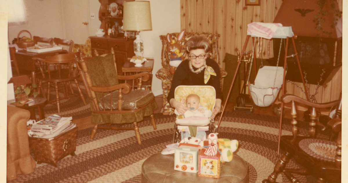 A faded color print from the early 1970s: Bill's grandmother Nanny, in glasses and a scarf, leaning over baby Bill in his walker in the living room of their old house, with blocks on the ottoman, a braided rug, and wood paneling.