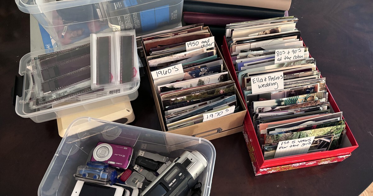 Boxes of family prints filed upright with handwritten decade labels, next to bins of old cameras, phones, film negatives, and discs on a dining table.